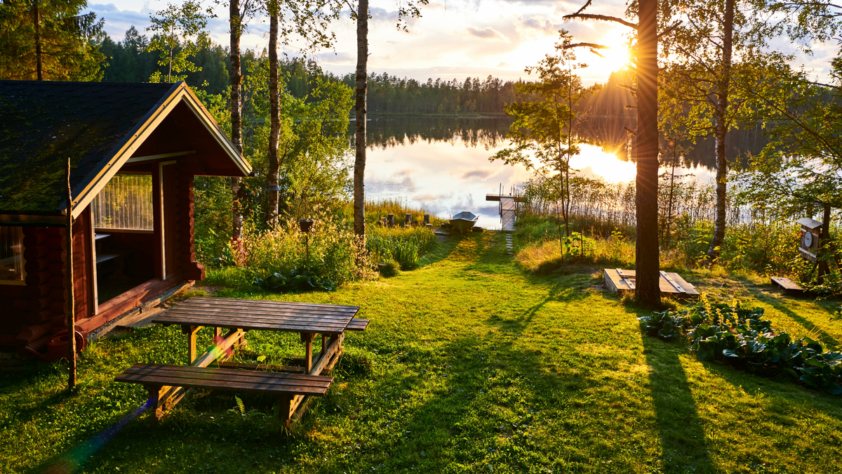 Aurinkoinen mökkipihamaisema, jossa näkyy järvi, laituri, soutuvene, puinen pöytä sekä punainen hirsirakennus
