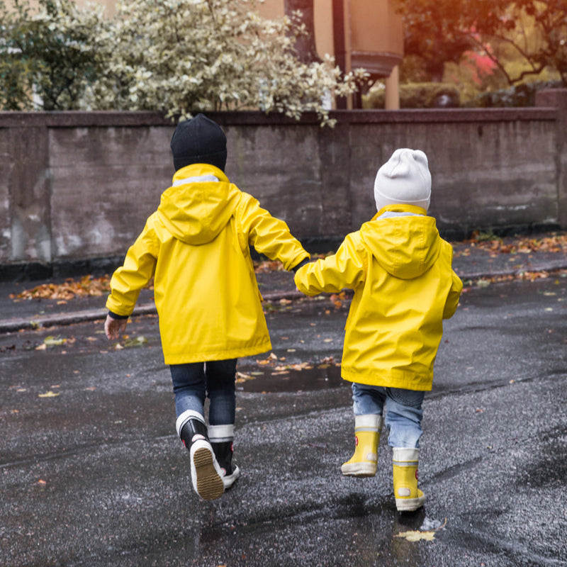Kaksi lasta keltaisissa sadetakeissa ja mustisa ja keltaisissa Hai Kids kumisaappaissa ylittämässä katua käsikädessään